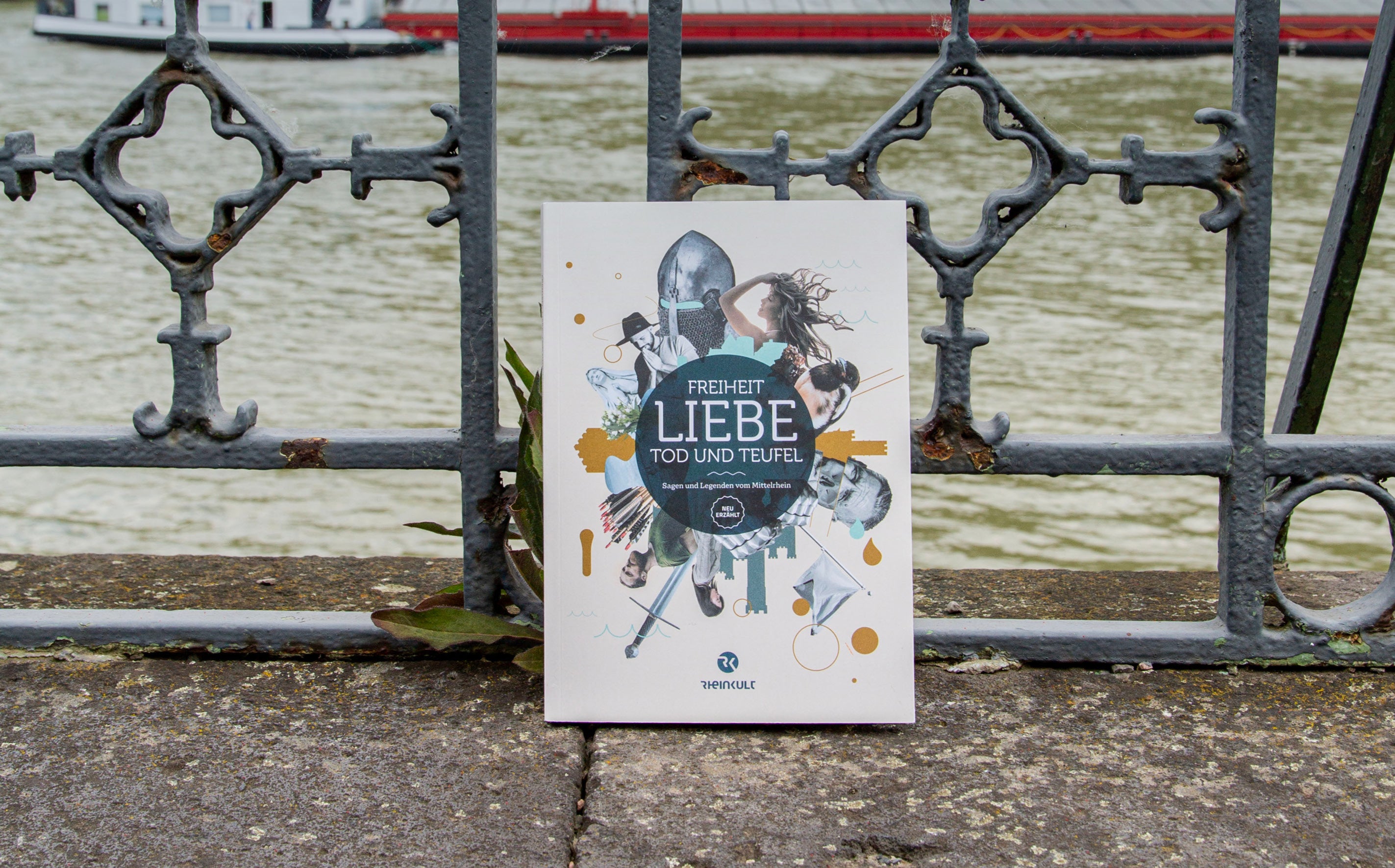 Das Rheinkult Sagenbuch „Freiheit, Liebe, Tod und Teufel“ lehnt an einem alten schmiedeeisernen mit Verzierungen. Im Hintergrund fließt der Rhein.