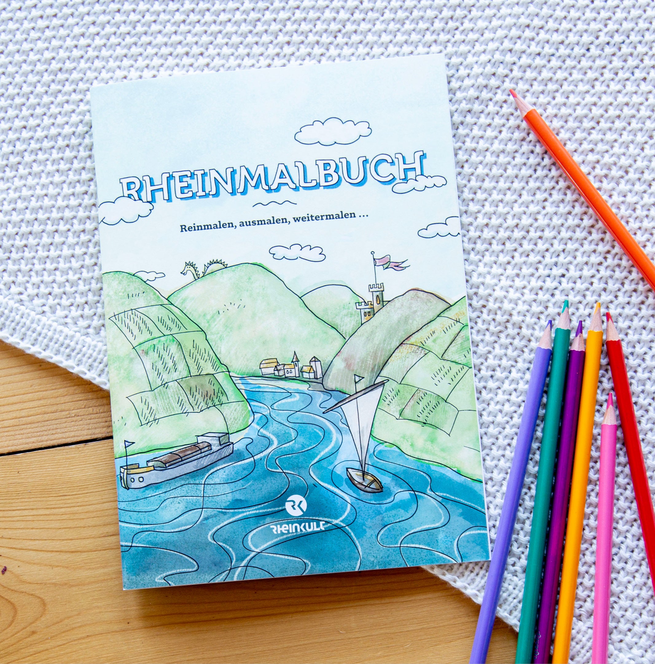 Auf einer Decke liegt das Rheinmalbuch der Marke Rheinkult neben einigen farbigen Buntstiften.
