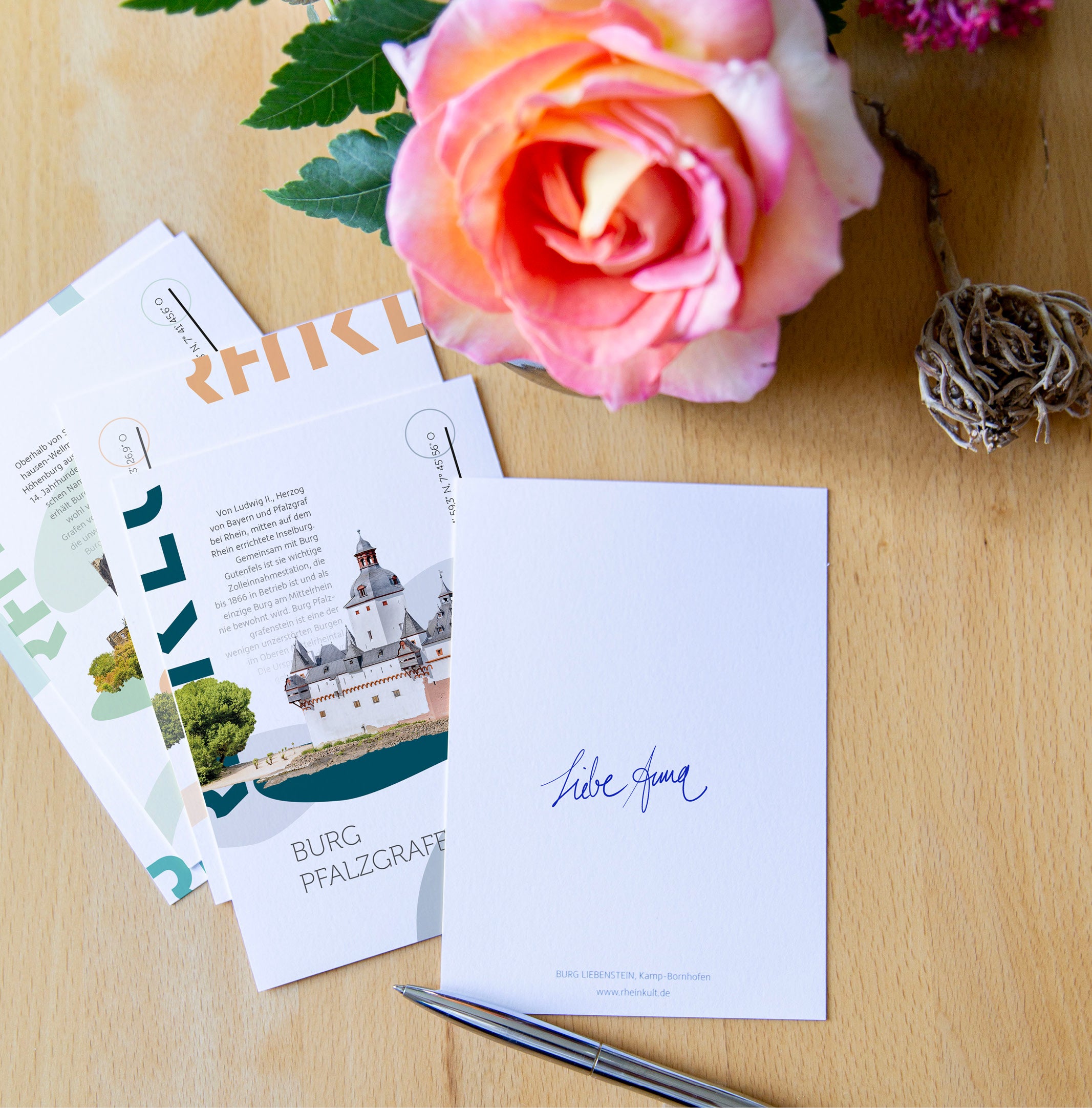 Rheinkult Postkarten, u. a. mit dem Motiv der Pfalzgrafenstein in Kaub, und eine Rose liegen auf einem Holztisch.