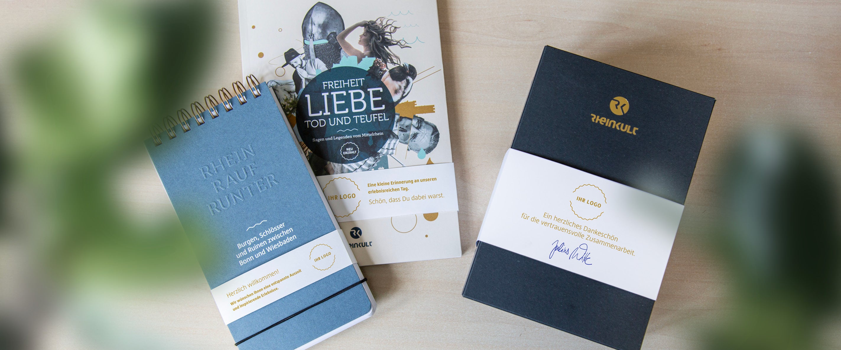 Drei Rheinkult-Produkte mit Banderolen als Individualisierungsvorschlag liegen auf einem Tisch: Der Spiralblock „Burgen, Schlösser und Ruinen zwischen Bonn und Wiesbaden“, das Sagenbuch „Freiheit, Liebe, Tod und Teufel“ und ein Betonuntersetzer-Set.