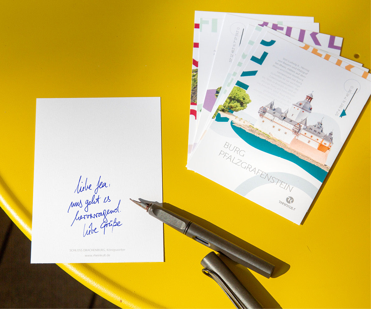 Auf einem gelben Tisch liegen auf einem kleinen Stapel Rheinkult-Postkarten. Daneben ein Füller und eine Karte mit beschriebener Rückseite: Liebe Lea, uns geht es hervorragend. Liebe Grüße. 