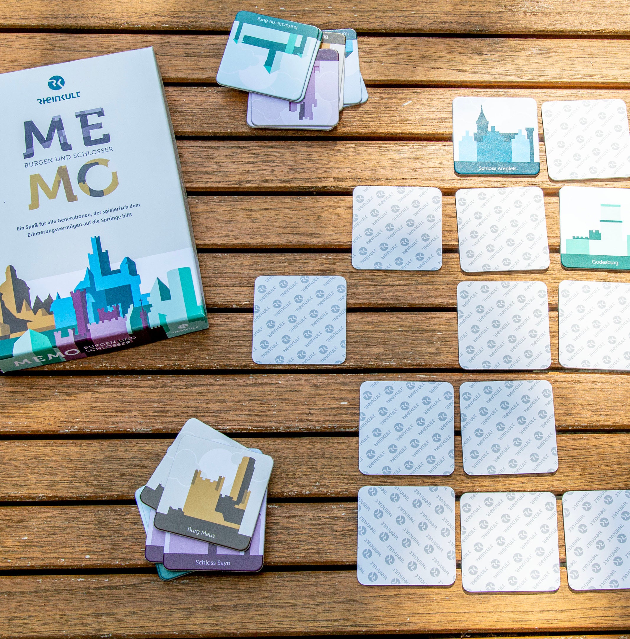 Karten des Rheinkult Memo-Spiels liegen teilweise verdeckt, teilweise gestapelt auf einem Holztisch neben der Verpackung.