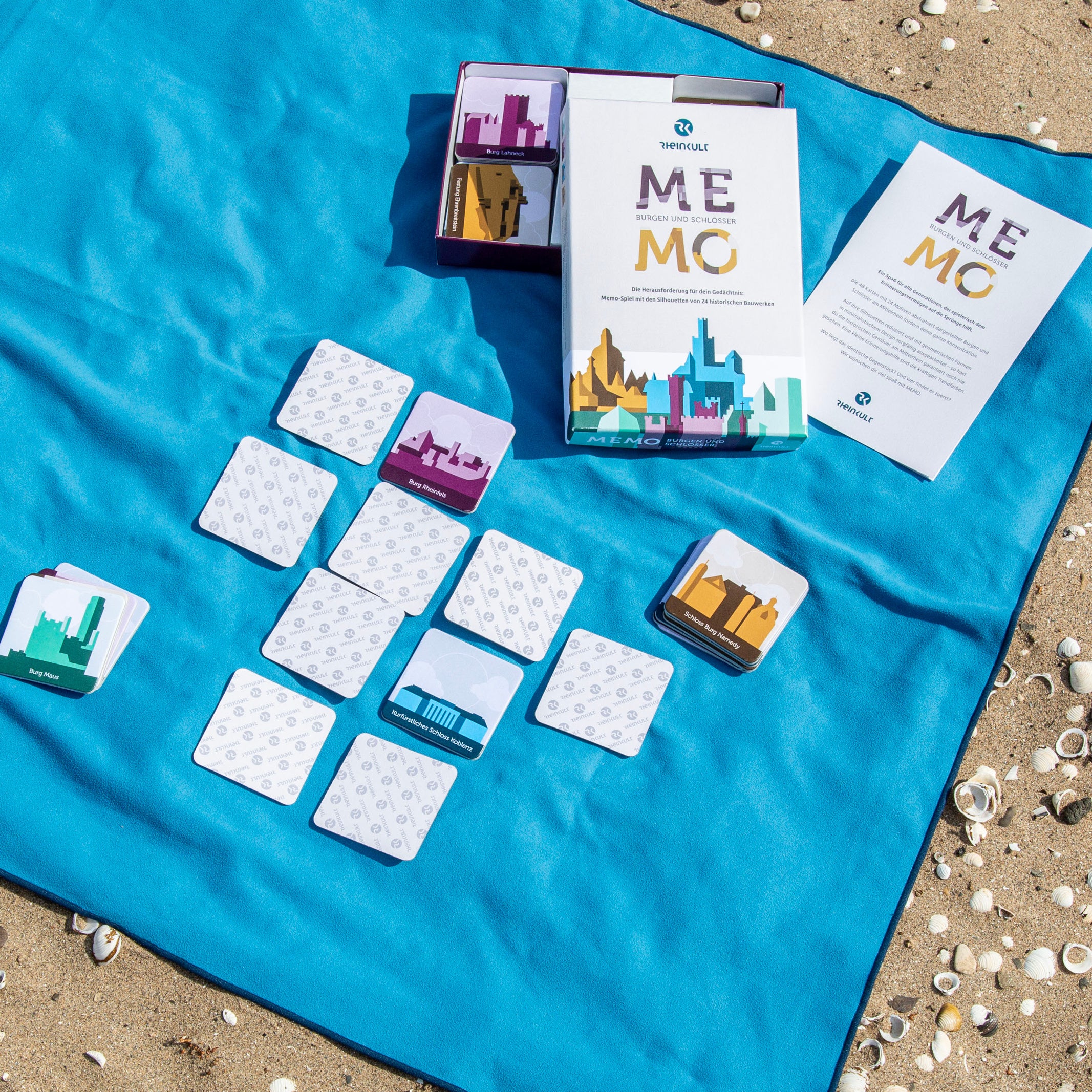 Das „MEMO Burgen und Schlösser“ zum Mittelrhein liegt auf einer blauen Decke an einem Sandstrand. Neben der geöffneten Verpackung liegen einige Spielkarten und ein Heft mit Kurzinfos zur Historie und Lage der im MEMO-Spiel gezeigten Burgen und Schlösser.