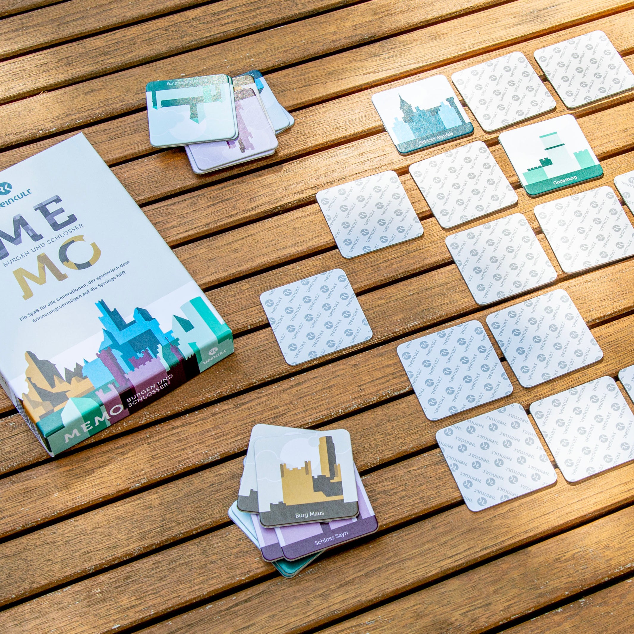 Auf einem Holztisch liegt das Rheinkult-Memo Erinnerungsspiel mit der Verpackung und kleinen Stapeln mit den Spielkarten. Darauf zu sehen sind die grafischen Darstellungen von Burgen und Schlössern am Mittelrhein.