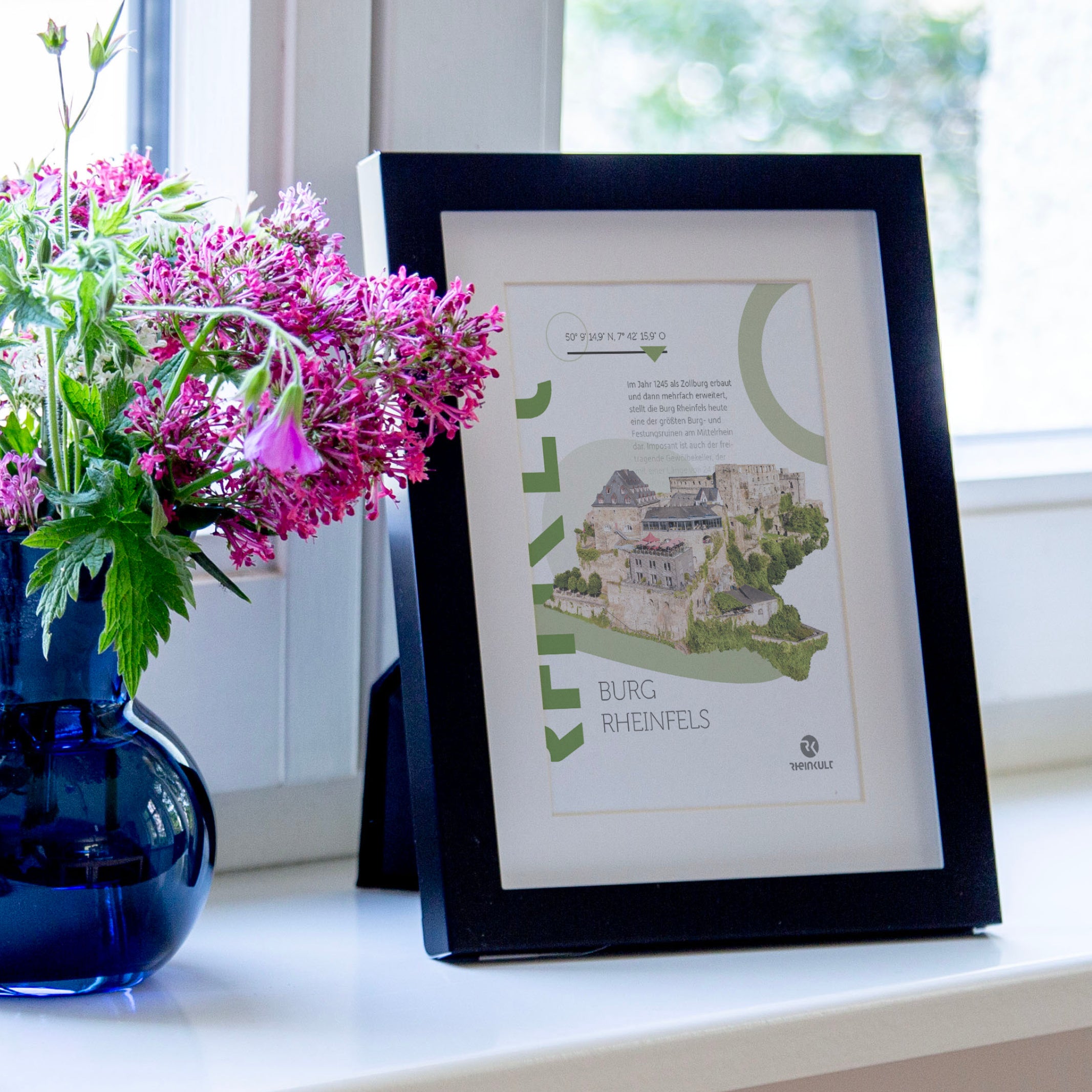 Auf einer Fensterbank steht ein Bilderrahmen mit der Postkarte „Burg Rheinfels“ neben einem kleinen Blumenstrauß.