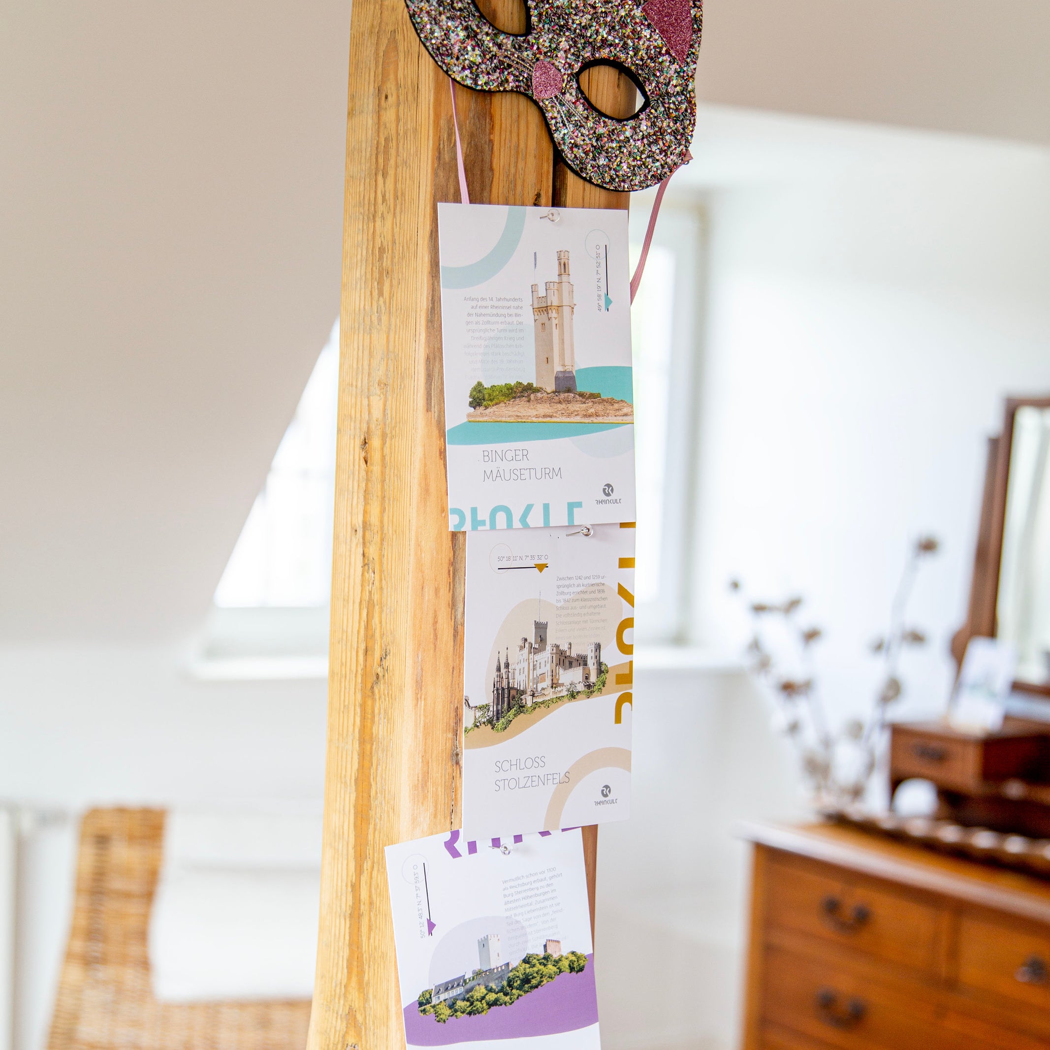 Postkarten der Marke Rheinkult hängen dekorativ an einem Holzbalken, befestigt mit Pins. Im Hintergrund ist ein Fenster und eine Kommode zu sehen. 