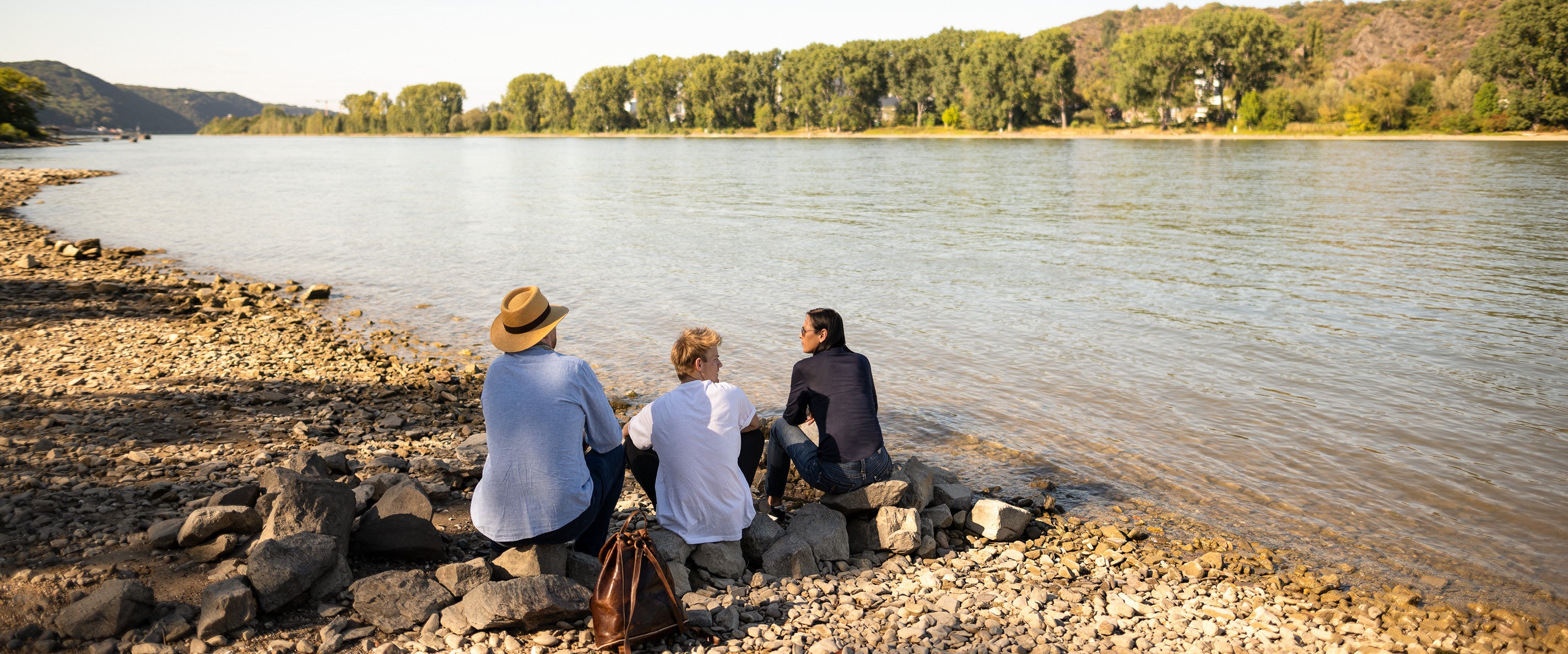 Das Team von Rheinkult sitzt auf Steinen am Ufer des Rheins. 