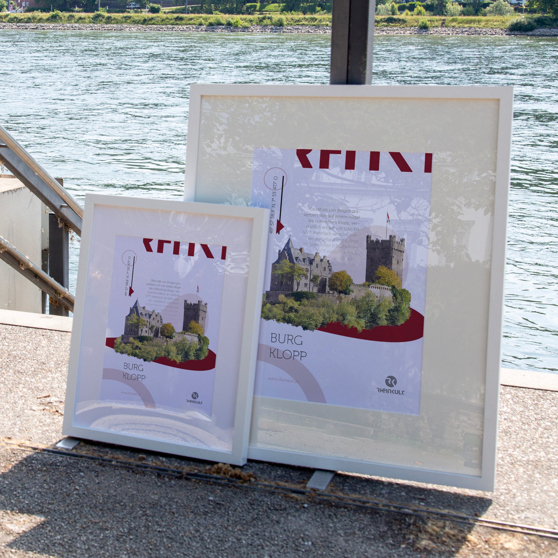 Zwei unterschiedlich große Rahmen stehen an ein Geländer gelehnt mit dem Rhein im Hintergrund. Sie zeigen das Rheinkult Poster „Burg Klopp“ in den Größen DIN A3 und DIN A4.