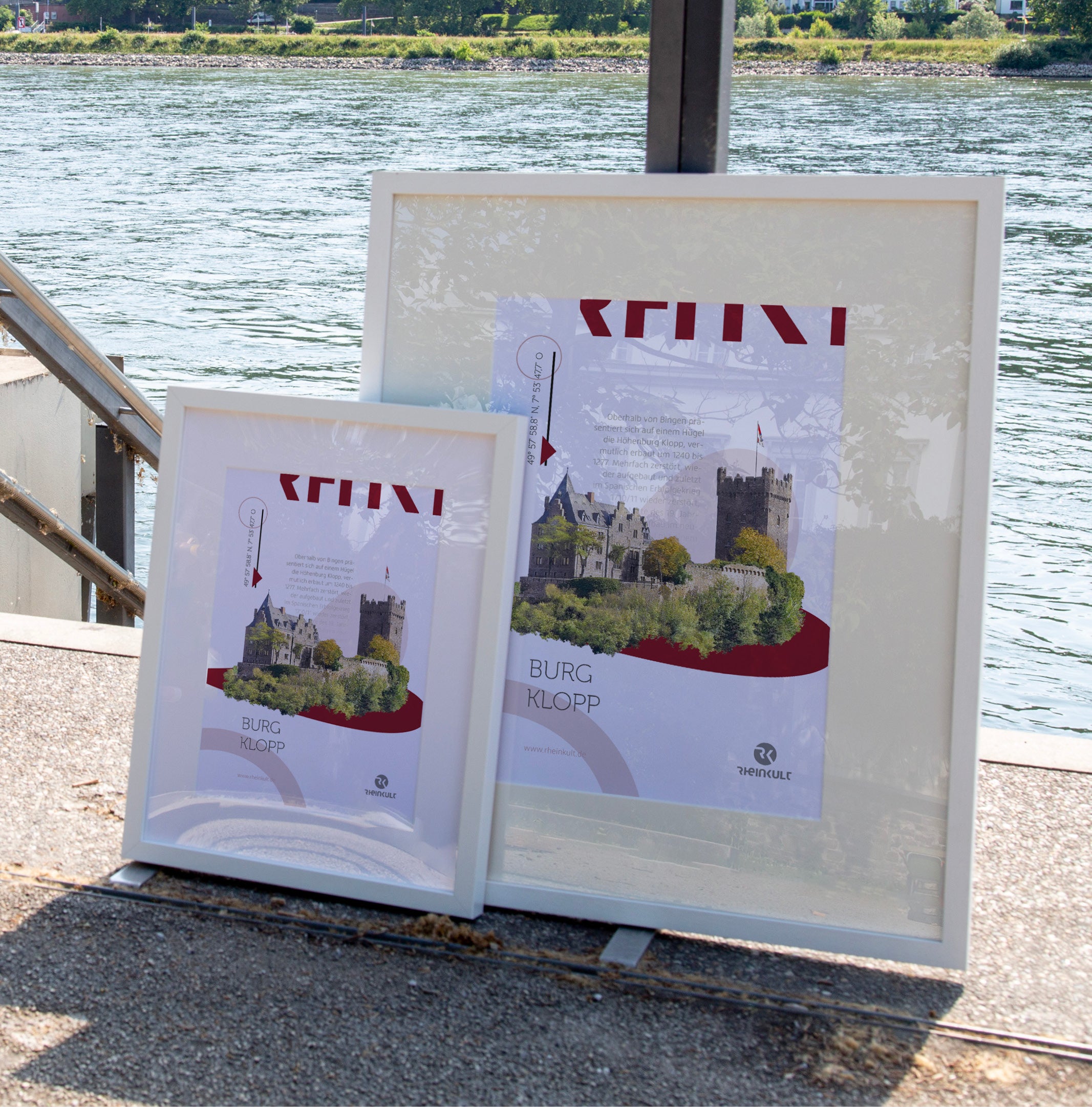 Zwei unterschiedlich große Rahmen stehen an ein Geländer gelehnt mit dem Rhein im Hintergrund. Sie zeigen das Rheinkult Poster „Burg Klopp“ in den Größen DIN A3 und DIN A4.