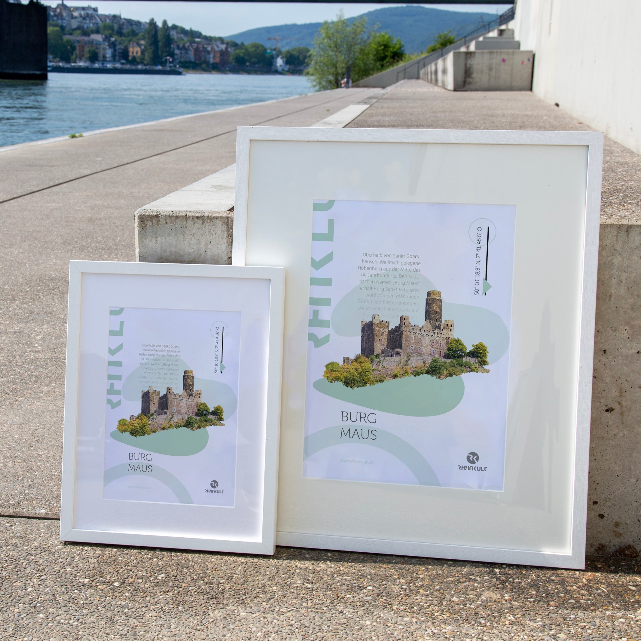 Zwei unterschiedlich große Rahmen stehen an eine Treppenstufe gelehnt mit dem Rhein im Hintergrund. Sie zeigen das Rheinkult Poster „Burg Maus“ in den Größen DIN A3 und DIN A4.