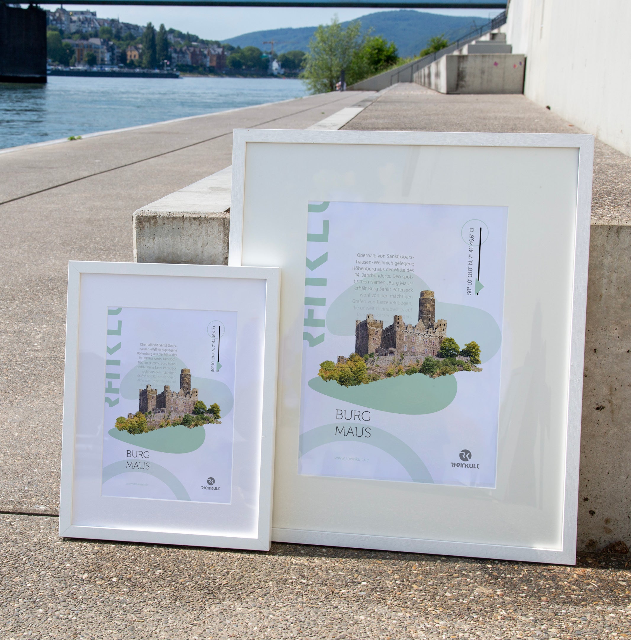 Zwei unterschiedlich große Rahmen stehen an eine Treppenstufe gelehnt mit dem Rhein im Hintergrund. Sie zeigen das Rheinkult Poster „Burg Maus“ in den Größen DIN A3 und DIN A4.