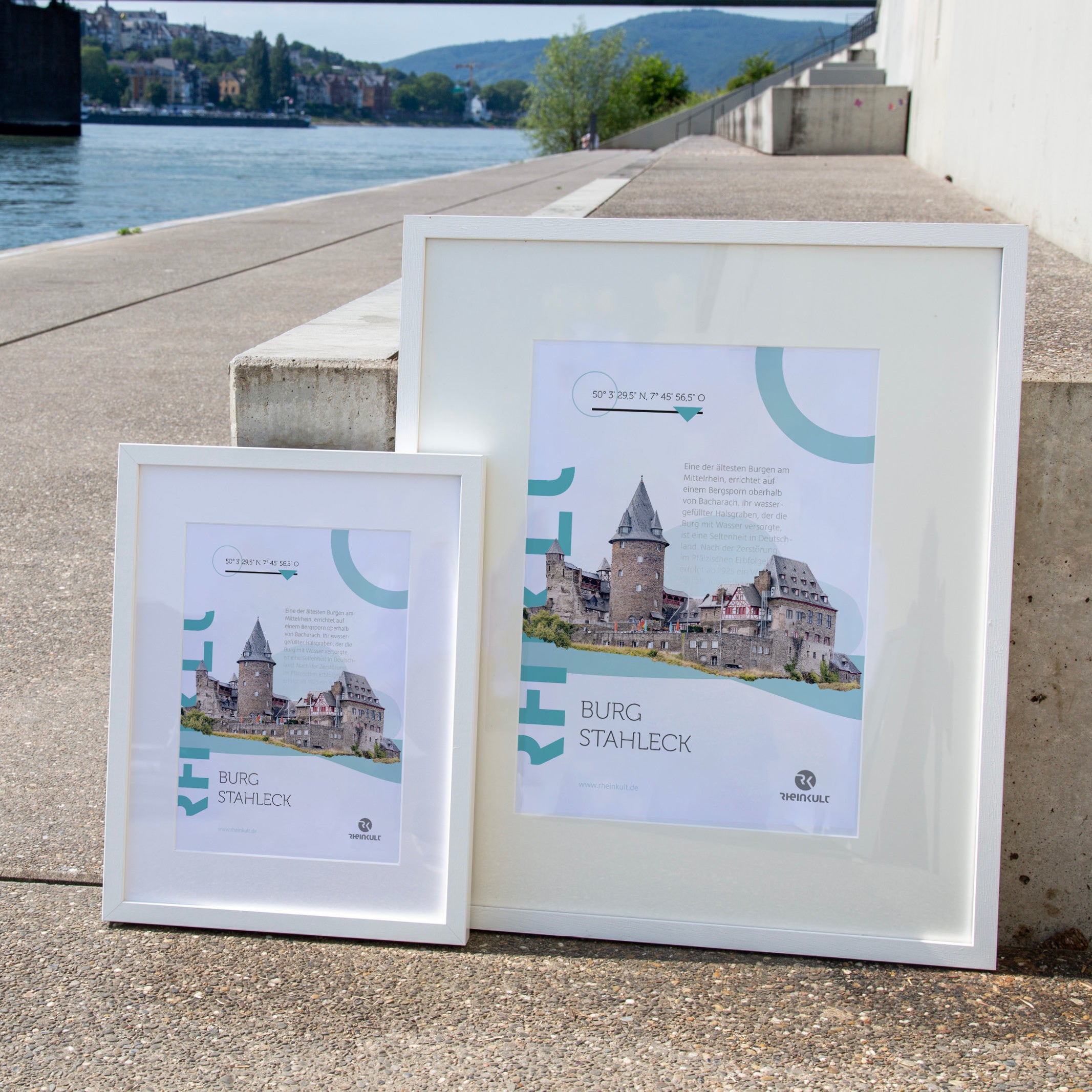 Zwei unterschiedlich große Rahmen stehen an eine Treppenstufe gelehnt mit dem Rhein im Hintergrund. Sie zeigen das Rheinkult Poster „Burg Stahleck“ in den Größen DIN A3 und DIN A4.
