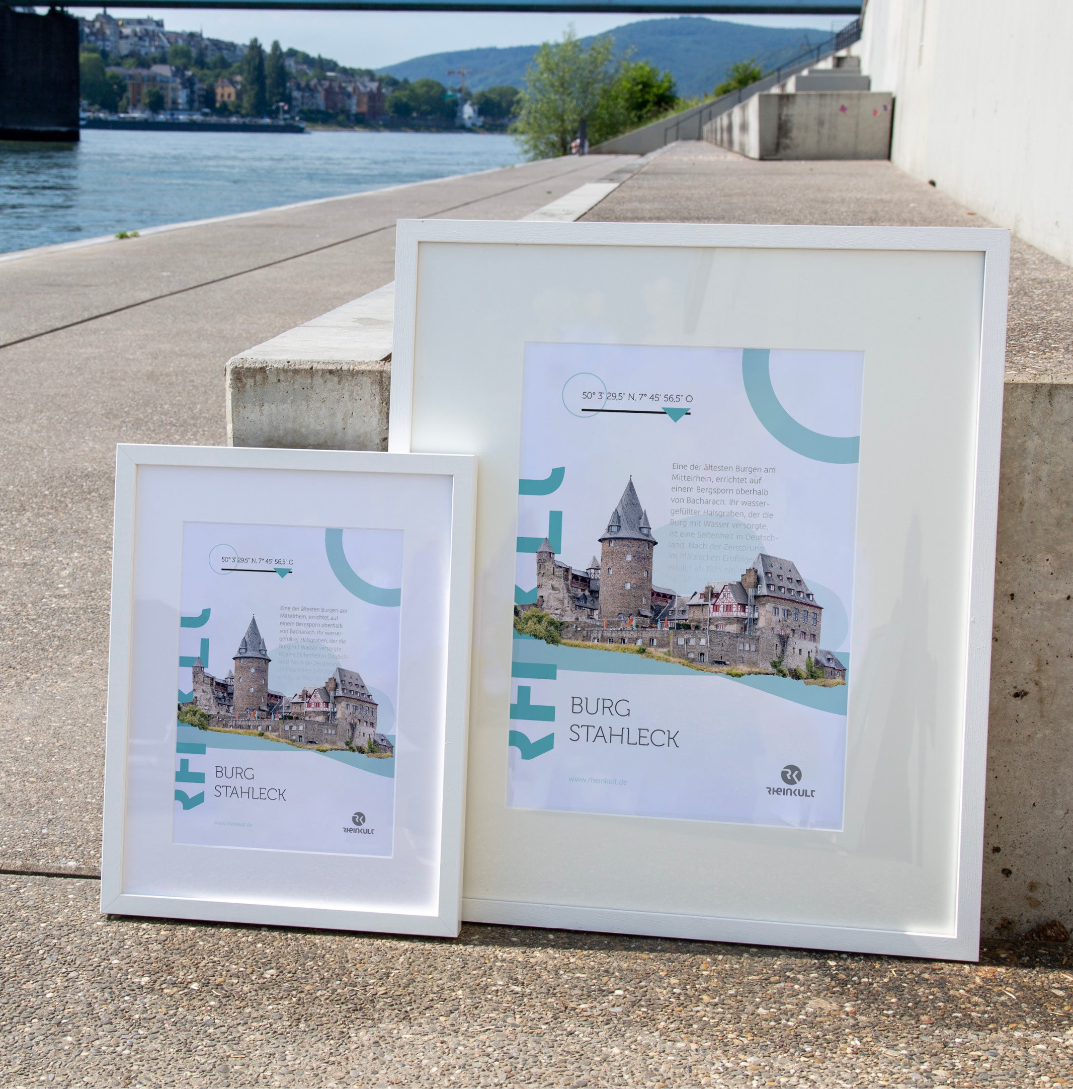 Zwei unterschiedlich große Rahmen stehen an eine Treppenstufe gelehnt mit dem Rhein im Hintergrund. Sie zeigen das Rheinkult Poster „Burg Stahleck“ in den Größen DIN A3 und DIN A4.