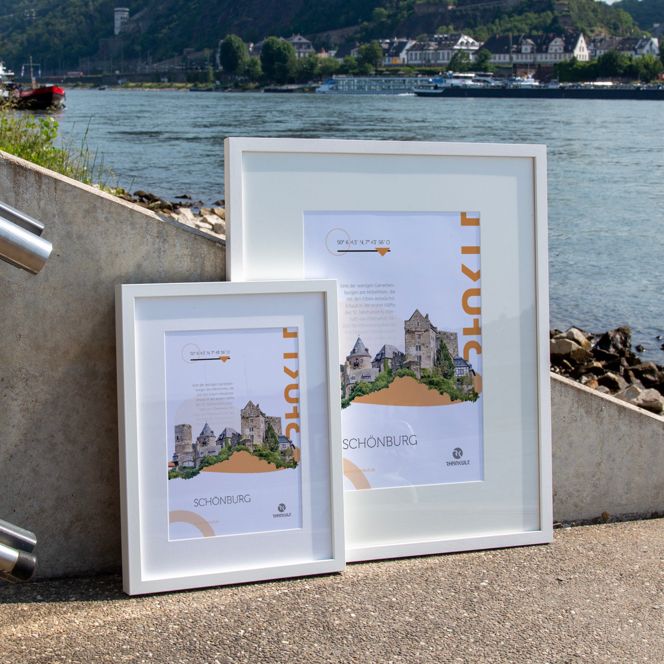 Zwei unterschiedlich große Rahmen stehen auf dem Boden vor einer Betonbrüstung mit dem Rhein im Hintergrund. Sie zeigen das Rheinkult Poster „Schönburg“ in den Größen DIN A3 und DIN A4.